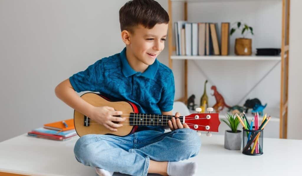 Smallest Instrument In The World A Boy Sitting On A Table, Playing A Small Ukulele