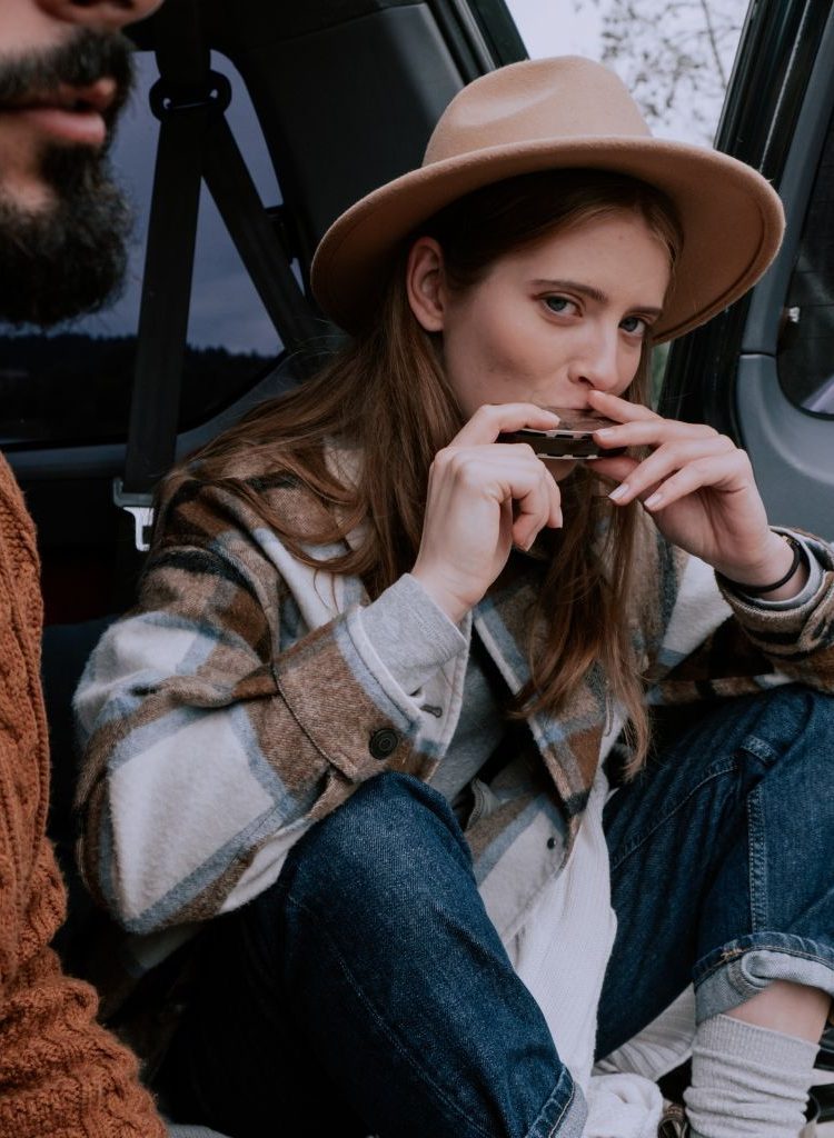 A Lady Wearing A Brown Hat Playing The Harmonica While Sitting On The Back Of A Car With A Friend A Lady Wearing A Brown Hat Playing The Harmonica While Sitting On The Back Of A Car With A Friend